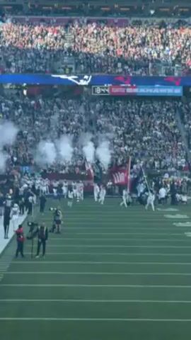 The video shows a large American football stadium filled with spectators, with a football team running onto the field through white smoke, waving flags, followed by staff and photographers. The field is green, and the atmosphere is energetic. The scoreboard shows a game between the Seattle Seahawks and another team, with "Patriots" visible on the scoreboard.