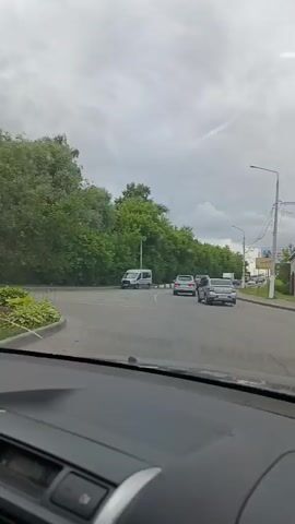 This media file captures a view from inside a car, looking out through the windshield. The scene is an overcast day, likely in the morning or afternoon, with a cloudy sky. The car appears to be driving on a road in a semi-urban or suburban area, with trees lining the left side of the road and buildings and a billboard visible on the right.

The main subjects are the road ahead and the surrounding environment. The road is asphalt and appears to be a two-lane road with some traffic. Several cars, including a silver sedan, a white van, and a white box truck, are visible on the road. There are also streetlights and power lines along the right side of the road.

The car's dashboard is partially visible in the foreground, suggesting the perspective of someone driving or being driven. The windshield has some visible cracks, which are a notable detail.

The setting includes lush green trees, suggesting a temperate climate, and a billboard with Russian text that is partially obscured and unreadable. The overall atmosphere is neutral, typical of everyday driving. The visible text on the billboard is difficult to discern, but the location context indicates Orekhovo-Zuyevo, Russia. The activity is simply traveling by car on a road.