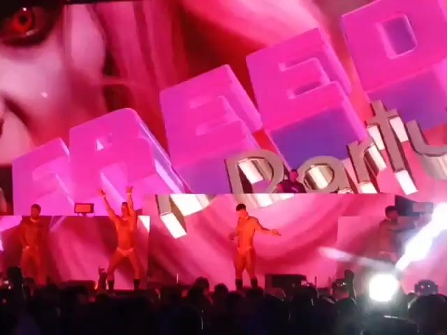 A crowded audience watches a stage performance. The performers include a DJ positioned above and behind five dancers in tan outfits. The stage displays "THE HOUSE OF X" in white letters above large, illuminated abstract shapes with pink and white hues.  Fire shoots upwards intermittently from behind the stage. Bright spotlights shine onto the performers and into the audience. Many audience members are holding up their phones, likely recording the performance.
