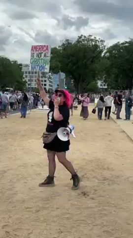 A woman with bright pink hair, wearing a black t-shirt, black shorts or a skirt, fishnet stockings, and dark boots, stands in the foreground, speaking into a white megaphone. She holds a sign above her head that reads "AMERICA IS NOT A CHRISTIAN NATION" in colorful, handwritten letters. She also has a leopard-print fanny pack slung across her chest. The ground is a light sandy or dirt color. In the background, many people are gathered, some walking, suggesting a protest or public event. Trees and modern buildings are visible further in the background under a cloudy sky. The overall atmosphere appears to be that of an outdoor demonstration.