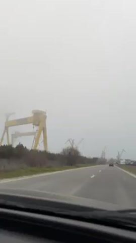 The video is taken from inside a moving car, driving on a road. The sky is overcast and foggy. On the side of the road are large yellow cranes. There is sparse vegetation growing along the edge of the road. A car is visible ahead in the distance.