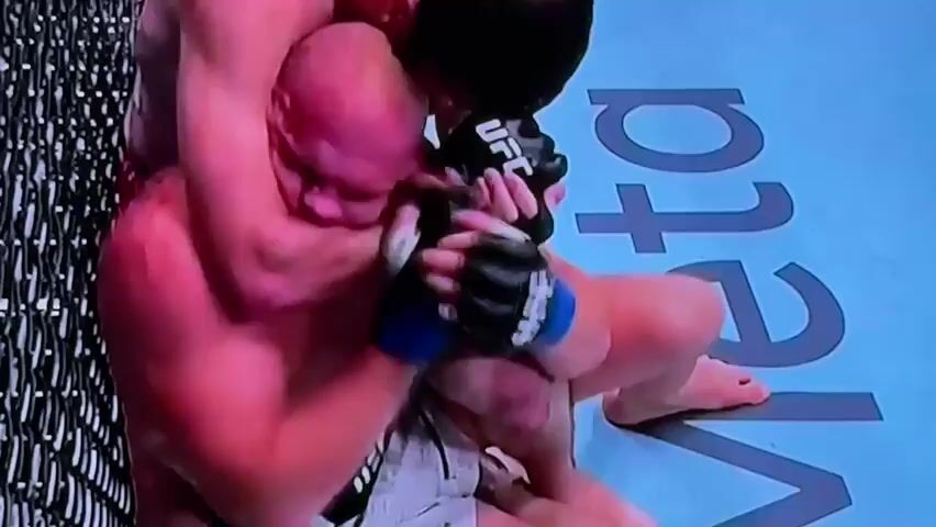 The video shows two men wrestling in what appears to be a mixed martial arts match. One man, who is bald and has a large build, is on his knees, with his back to a chain-link fence. The other man, who is taller and has dark hair, is behind him, wrapping his arms around the bald man's torso. The taller man's hands are clasped around the bald man's chest, and he appears to be trying to lift or control him. Both men are wearing MMA gloves. The background is a blue mat with white lettering that appears to say "UFC." The lighting is bright and pinkish, suggesting it may be from overhead lights. There is some red staining on the bald man's chest, possibly blood. The camera is positioned above and to the side, looking down at the two men.