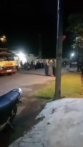 A nighttime outdoor scene shows a group of people, some wearing headscarves, standing near a yellow dump truck. There are also other vehicles present, including a white dump truck and motorcycles parked in the foreground. The area appears to be a street or parking lot, with a pole and some greenery visible. The lighting suggests artificial light sources are illuminating the scene.