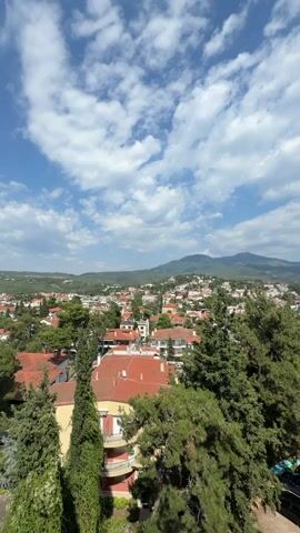 This is a vertical, wide-angle shot of a residential town nestled in a valley, with rolling hills and mountains in the background. The sky is a vibrant blue with scattered white clouds. The town is characterized by red-tiled roofs, interspersed with lush green trees. In the foreground, several tall, mature evergreen trees dominate the scene, with one particularly large tree to the right casting a shadow on a dirt parking area.

The parking area contains several cars, including a dark SUV and a few smaller vehicles. A multi-story building with balconies is visible on the left side of the frame, also featuring red-tiled roofs and balconies. The overall impression is one of a peaceful, suburban or small-town setting. The time of day appears to be late morning or early afternoon, given the bright sunlight and clear skies, suggesting good weather. The location appears to be in a hilly or mountainous region, consistent with the Panorama Municipal Unit in Greece. No people are directly visible in the frame, and no text is present. The scene evokes a sense of calm and natural beauty.