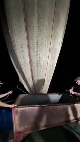 The video shows two men, one on the left and one on the right, holding ropes attached to a large, funnel-shaped net. The net is being lowered into a long, rectangular metal container, which appears to be a bin or trough. The scene is dimly lit, suggesting it is nighttime, and the background is dark. The men are focused on their task, and the net is in the process of being emptied into the container. The metal container shows signs of rust and wear.