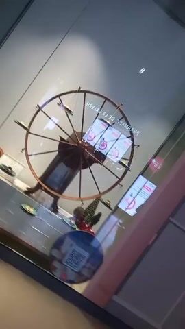 The video shows a shop window display featuring a large, antique-looking scale balanced like a Ferris wheel. The scale has multiple pans hanging from its circumference, each holding a small, possibly decorative, item.  The display also includes small Christmas trees, decorative objects resembling pine cones, and promotional materials. A QR code is visible on a sticker. The scene appears to be inside a shopping mall or commercial building, given the clean tile flooring and the presence of the shop window. The camera moves slightly, offering different views of the display.