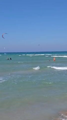 The video shows a bright, sunny day at a beach with clear blue skies. The ocean is turquoise and waves are breaking on the shore. There are several people visible in the water, some swimming and others engaged in watersports. A kitesurfer is visible in the distance, with their kite in the air. Another person on a board is also in the water. The beach itself is sandy and appears to be a popular spot for recreation. The water is calm with moderate waves.
