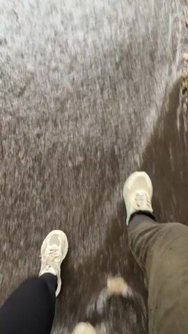 The video shows a first-person perspective of someone walking through a flooded area, possibly a sidewalk or street. Their feet, wearing light-colored sneakers and dark leggings, are visible at the bottom of the frame. As they walk, their feet splash through the water, creating ripples and splashes. In the background, the water appears murky and covers a wide area, with some fallen leaves visible on the right side. The action is a series of steps taken through the water, with each step causing a more pronounced splash.