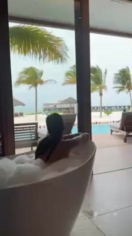 A woman with long dark hair sits in a white freestanding bathtub filled with bubbles. She faces away from the camera, looking out through a large glass door. Outside, there is a swimming pool, lounge chairs, palm trees, a sandy beach, and an ocean view. A man in a green shirt and black pants walks past, holding a long-handled tool, likely for pool maintenance. The weather appears overcast.  The room's interior is modern, with tiled floors. The tub appears to be next to the glass door, offering a view of the tropical scenery.