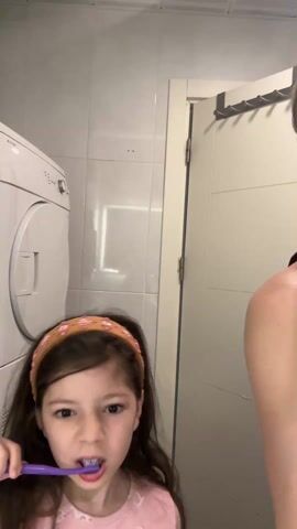 In a well-lit bathroom, a young girl with long, dark hair and a patterned headband is brushing her teeth. She wears a light-colored top and uses a purple toothbrush, making some animated facial expressions as she brushes. To the right, partially visible, is an adult woman, whose head is mostly bowed, showing her dark top and hair with some lighter streaks.

The background features white tiled walls. On the left, a white washing machine or dryer unit is visible. To the right of the frame, a light-colored door is partially open, with a rack of hooks hanging on its inner side. A rectangular reflection, possibly from a light fixture, can be seen on the tiled wall between the washing machine and the door. The overall scene suggests a routine activity, likely morning or evening, with the girl actively engaged in dental hygiene.