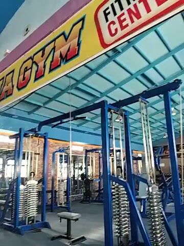 The video shows the interior of a gym with blue-colored weight machines. A sign above the gym reads "Fit & Fun Gym". There are no people visible in the gym. The gym appears to be well-equipped with various weight machines and free weights. The floor is a dark blue color, and the ceiling is also blue with white lights. The walls are a light brown brick pattern.