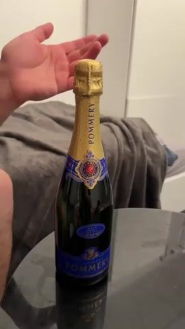 A medium shot shows a person attempting to open a bottle of Pommery Brut Royal champagne while seated at a small black table. The bottle, dark green with a gold foil-wrapped neck, is positioned vertically on the table. The person’s hands, light-skinned with visible veins, are engaged in manipulating the foil and attempting to twist the bottle’s neck. In the background, out of focus, is a bed with a gray blanket and the corner of a white cabinet. The room appears to be a bedroom or small apartment.