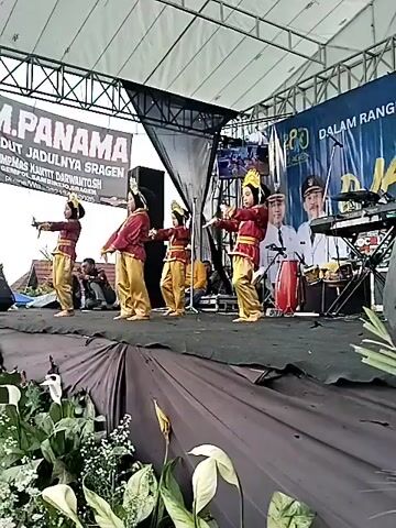 Here is a description of the video:
The video shows four young dancers performing on a stage outdoors. They are dressed in matching traditional costumes consisting of a maroon top, golden pants, and headpieces. The stage is covered in brown fabric, with floral decorations at the front. Behind the dancers are musical instruments, including a keyboard and drums, with a large banner in the background featuring text and images of men in uniform. Several individuals are seated in the background, observing the performance. The stage is under a white tent-like structure, supported by a metal frame.