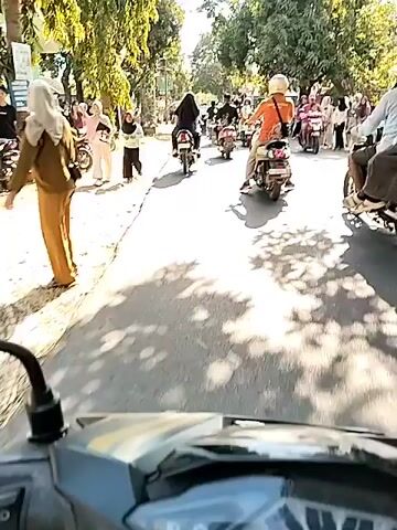 The video captures a view from a motorcycle traveling down a paved road. On the left side of the road, there is a dry, grassy embankment with several parked motorcycles and some people standing. Further down the left side, there are buildings with green walls and trees. The road itself has a rough texture and is partially covered in the shadows of trees. On the right side of the road, there are more trees casting shadows onto the asphalt. Several motorcycles are in front of the camera, moving in the same direction. The people on these motorcycles are wearing helmets and varying clothing, including a person in an orange shirt and a person in a light-colored shirt. The overall environment appears to be a sunny day in a rural or suburban area with a moderate amount of traffic.