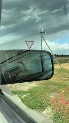 The video shows a view from inside a vehicle, looking out the passenger side window and in the side mirror. The side mirror reflects the interior of the car, including a cell phone and a steering wheel. Outside the car, a road is visible, with a ditch and grass on the side. In the distance, there is a field and a road with a red car driving on it. There are also traffic signs visible, including a yield sign and a speed limit sign indicating 60. The sky is cloudy. The vehicle appears to be moving.