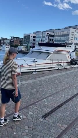 The video shows a scenic waterfront in Karlstad, Sweden. In the foreground, three individuals are walking along a cobblestone path beside a canal. The first person, from behind, is wearing a white t-shirt and a beige skirt, holding a black jacket. The second person is wearing a white t-shirt and sunglasses, with blonde hair. The third person, also from behind, is wearing a striped t-shirt, shorts, and a baseball cap. They appear to be enjoying a leisurely stroll on a sunny day.

The canal is filled with blue water, and several boats are docked along the waterfront. The boats range from smaller vessels to a large, white yacht with "PRINCESS 50" visible on its side. In the background, modern buildings with large windows line the canal, suggesting an urban or commercial area. There is a prominent tower structure with a viewing platform visible further down the waterway. A sign with a red circle and a diagonal line through a car and a motorcycle symbol, indicating "no vehicles," is also present near the boats. The sky is blue with scattered white clouds, and the overall atmosphere seems peaceful and pleasant, indicating it's likely late afternoon or early evening. The lighting suggests the sun is low in the sky, casting a warm glow.