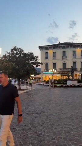 The video shows a waterfront promenade at dusk. In the foreground, a man in a black polo shirt and light-colored shorts walks from left to right. Behind him, a woman in a blue top and patterned skirt also walks from left to right. Further in the background, several people are strolling along the promenade, and some are seated on benches. A large tree is visible on the left side of the frame. The promenade is paved with cobblestones. Across the water, hills are visible. There are streetlights and buildings along the promenade. Boats are docked in the water. The sky is a clear blue with some clouds. The overall atmosphere appears to be peaceful and leisurely.