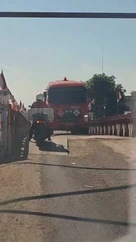 A red fuel tanker truck with "ELNUSA PETROFIN" printed on the front drives across a bridge. A motorcycle with a rider wearing a white helmet is in front of the truck, also on the bridge. The bridge has red barriers and flags on either side. The sky is blue and clear. The truck has hazard symbols on the front, indicating it is carrying dangerous materials.