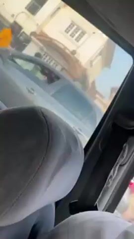 From inside a car, the video shows four men gathered around the back of a silver car.  One man wears a pink polo shirt with the number nine on the back, another wears a gray T-shirt with the number 78 on the back. Another man wears a white T-shirt and the fourth man wears a black shirt. They appear to be conversing.  A yellow building with a window and gray roof is in the background.  The video is shaky and shows a gray car seat in the foreground.