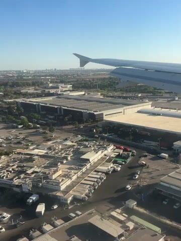 This media file captures an aerial view from an airplane window overlooking a bustling airport or industrial complex. The sun is high in the sky, suggesting it is midday, and the weather appears clear with a bright blue sky.

The primary subjects are multiple airplanes parked on the tarmac, with their wings and engines clearly visible. In the foreground, there are various buildings with flat, light-colored roofs, some featuring a distinctive red and white checkered pattern. Surrounding these buildings are numerous trucks, cargo containers, and other industrial equipment, indicating a logistics or cargo hub. The overall scene depicts a busy operational area, likely an airport's cargo terminal or a large industrial zone.

Notable details include the airplane wing in the upper right portion of the frame, providing context for the aerial perspective. The landscape beyond the immediate airport area shows a city skyline in the distance, hinting at the urban environment.

Visible text includes "Challenge Accepted" printed on the side of one of the airplanes.

The media evokes a sense of activity and scale, showcasing the logistical operations of air travel and freight. The clear weather and bright sunlight contribute to a feeling of a productive day. The location cues, combined with the visual elements, suggest this scene is likely in Zeitan, Israel, an area known for its industrial zones and proximity to transportation infrastructure.