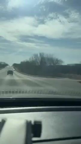 The view from inside a car driving on a highway under a partly cloudy sky is captured. Other vehicles are present on the road. Leafless trees line the side of the highway. The dashboard of the car is visible in the foreground.