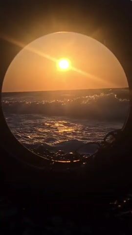 A wide circular frame, possibly a tire, looks out onto a beach at sunset. The sun is a bright orange orb, low on the horizon, casting a golden glow over the water. Gentle waves roll in, their crests highlighted by the sun's rays. The foreground is dark and out of focus, suggesting the perspective is from within a shadowy object. Some dark, rope-like material is visible in the lower right corner of the frame.