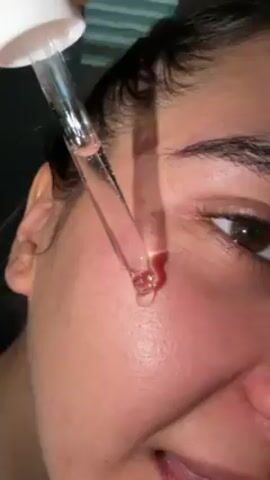 A close-up shot shows a smiling woman applying a clear liquid serum to her face using a dropper. The serum is dispensed from the dropper onto her cheek in small droplets. The woman has dark hair and brown eyes, and her skin has visible pores. The background is slightly blurred.