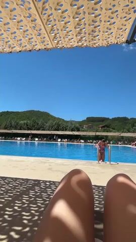 The video begins with a low-angle, first-person perspective, showing a person's legs resting on a lounge chair under a patterned sunshade. The sunshade casts a dappled shadow pattern on the foreground. Beyond the lounge chairs, a bright blue swimming pool is visible, bordered by a tiled deck. In the middle distance, people are seen lounging on chairs and walking around the pool area. A woman in a swimsuit is walking towards the pool ladder. Further back, a lush, green, tree-covered hillside forms the backdrop under a clear blue sky with a few scattered clouds. The scene appears to be a resort or a public pool on a sunny day.