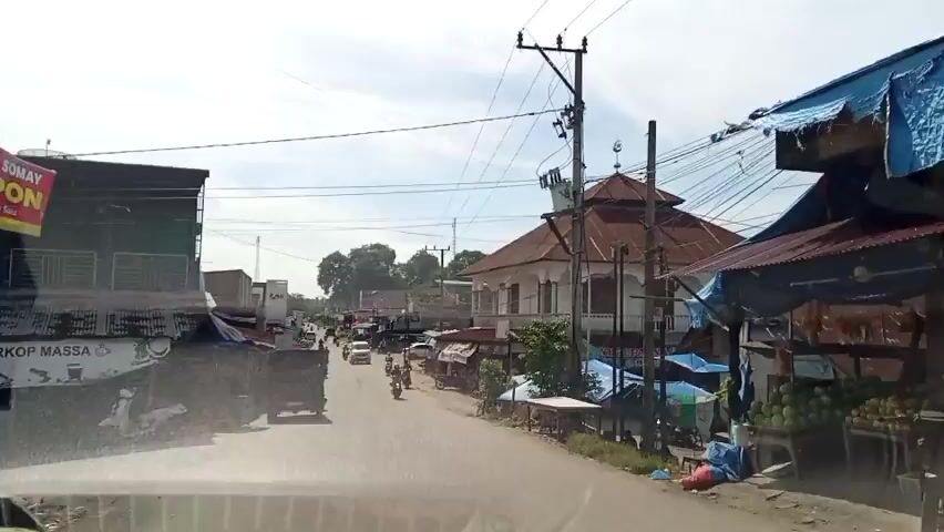 The video shows a street view from a moving vehicle. The street has two lanes of traffic. On the right, there are numerous stalls selling items, some covered by blue tarps. In the middle of the shot, a mosque with a red-tiled roof and a minaret is visible. Various power lines crisscross overhead. On the left, several buildings line the street. Vehicles, including motorcycles and cars, are traveling in both directions. The sky is partly cloudy.