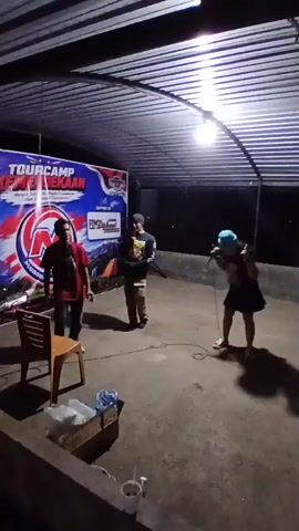 The video shows three people standing under a roof in what appears to be an outdoor setting at night. A banner with text, some of which is visible as "TOURCAMP KOTA KEBANGKAAN" and a logo with an "M", hangs behind the leftmost person. This person is wearing a red shirt and black pants, standing next to a plastic chair. The middle person is wearing a black t-shirt with a graphic and tan pants. The rightmost person is wearing a black t-shirt, dark shorts, and a light blue head covering, holding a microphone and dancing. A microphone is also visible on the ground near the middle person, and another microphone is held by the middle person. A light fixture hangs from the roof, illuminating the area. There are some boxes and cups on a ledge in the foreground.