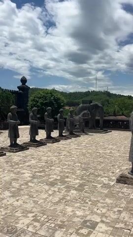 The video opens on a wide shot of an outdoor courtyard paved with square tiles. In the foreground, a row of weathered stone statues of human figures stand in profile, facing left. To the right of these, the perspective shifts to show another statue of a human figure in profile, facing right. Beyond the statues, a large, ornate stone archway stands prominently against a blue sky with scattered white clouds. In the distance, rolling green hills are visible. To the left of the archway, a stone sculpture of an elephant and possibly a chariot or carriage can be seen. Several tourists are standing near the archway, some holding umbrellas to shield themselves from the sun. The overall atmosphere is one of historical significance and tourist visitation.