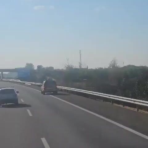 The video captures a view from inside a vehicle traveling on a highway. The camera is angled slightly upwards, showing the road ahead and the surrounding landscape. On the right side of the highway, there is a white guardrail. Beyond the guardrail, there is a dry, vegetated embankment with sparse trees and bushes. A utility pole with some antennas is visible in the distance on the embankment. On the left side of the highway, a blue road sign is visible, indicating distances to various locations, including "Alfajarin" at 4 km. A white car is driving in the left lane ahead of the camera. In the right lane, closer to the camera, a red pickup truck with a blue light bar on top is visible. The truck has "BOMBEROS" written on the back, indicating it is a fire department vehicle. The sky is clear with a few white clouds. The overall lighting suggests it is daytime.