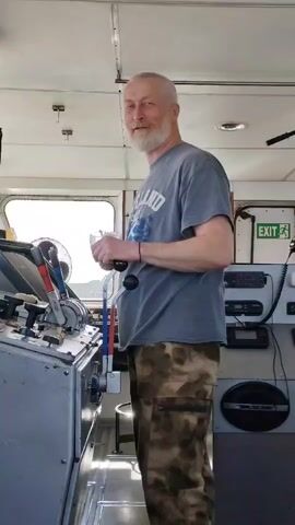 A medium shot captures a middle-aged man standing inside what appears to be the control room of a boat. The man, who is white, has a grey beard and short grey hair. He is wearing a grey t-shirt with the word "Finland" printed on it and camouflage pants. In his hands, he holds a small, dark-colored object. He looks towards the camera and smiles. To his left is a control panel with various levers and gauges. Behind the man, on the right side of the frame, there is a wall with an "EXIT" sign and some electronic equipment. A window is visible in the background, providing a view of a landscape. The room is brightly lit, and the walls and ceiling are white. The man is facing towards the camera and slightly to the left. He appears to be engaged in a conversation, as he is looking at the camera and speaking.