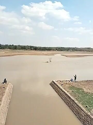 A wide shot shows a muddy river with dry, brown land on the far bank. Two people are in the water, one is on a floating object, and the other is next to him, holding a large, triangular fishing net. A person is sitting on the edge of a stone-lined embankment in the foreground. Another person is standing on the same embankment, holding a fishing rod. The sky is partly cloudy.