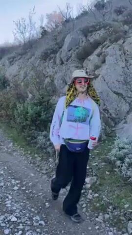 A person is walking uphill on a rocky path. They are wearing black pants, a white long-sleeved shirt with colorful graphics, a beige hat, red sunglasses, and a colorful fanny pack. They also have a backpack. The person is holding a can of soda. The terrain is rocky with sparse vegetation. The person says something in what sounds like Russian. The video ends with the person turning away from the camera.