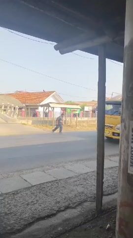 A yellow minibus with red and white striped decals is driving down a paved road. The door of the minibus is open, and two people, a man and a woman, are standing on the step, with the woman appearing to board or disembark. The man is holding onto the side of the bus. The environment is a roadside with buildings and trees visible in the background. The sun is shining.