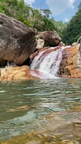 A low-angle shot shows a waterfall cascading over reddish-brown rocks into a body of clear green water. Large boulders and lush green foliage surround the waterfall. Two people are visible standing on a rock near the top of the waterfall.