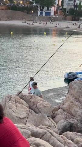 A girl in a white shirt and red pants walks along a concrete pier towards a man in a grey shirt and a baseball cap. The man is holding a fishing rod. In the background, a beach is visible with people relaxing and swimming. A few yellow buoys are scattered in the water. A small inflatable boat with an outboard motor is docked on the right side of the pier. The scene is lit by natural light, suggesting it is daytime. The foreground is dominated by large, textured rocks.