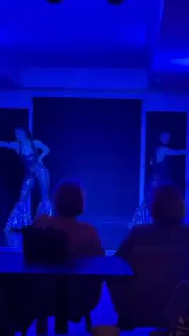 Two dancers in silver jumpsuits are performing on a small stage. The stage has a dark backdrop and is lit with colorful lights that change from purple to green to blue. Two audience members are visible, seated at a table. One of them is holding a cup. The lights change in coordination with the music.