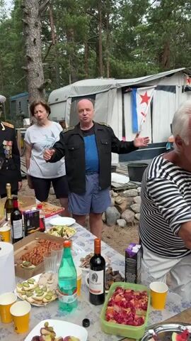 A group of people are gathered outdoors around a picnic table laden with food and drinks. A bald man in a black jacket and blue shorts stands in the center, holding a shot glass and gesturing with his hands. A woman in a white t-shirt stands to his left, also holding a small glass. To the right, a man in a striped shirt leans in, offering a toast. In the background, a camper trailer is visible with a flag bearing a red star. The scene appears to be a casual outdoor gathering with a celebratory atmosphere.