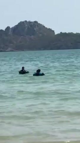 The video shows two people swimming in a body of water near a landmass. The water is light green and slightly choppy. One person is swimming without any flotation device, while the other appears to be using an inner tube. The landmass is covered in trees and brush. The sky is clear and bright.