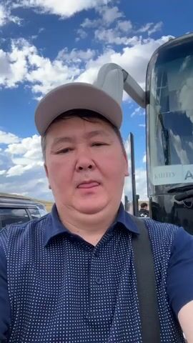 This media file is a selfie-style video featuring a man in a blue, polka-dot shirt and a grey baseball cap. He is standing in front of a large black bus, which appears to be a passenger coach. The bus has "СПУ" and "АБ" visible on its front, likely indicating a destination or company name. The man is looking directly at the camera with a neutral expression.

Behind the man and the bus, the scene suggests an outdoor setting. The sky is a bright blue with scattered white clouds, indicating it is daytime and the weather is fair. The ground appears to be dry and somewhat barren, with hints of rolling hills or flat terrain in the distance. There are other vehicles and people visible in the background, including what looks like a dark-colored SUV to the left of the frame and another person standing near the bus door.

The man's posture and the selfie framing suggest he is documenting his presence at this location, possibly before or after a journey. The overall impression is of a travel or waiting scenario, perhaps at a bus station or a stop along a route in a rural or semi-arid region. The location context of Bakty, Kazakhstan, aligns with this visual impression of open landscapes and transit.