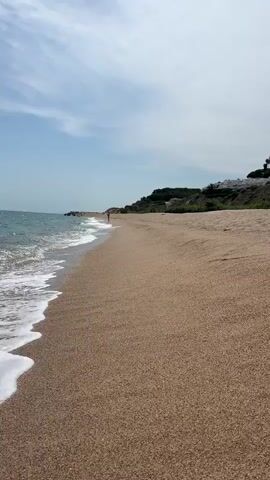 The video shows a beach with a shoreline and a person in the distance. The water is a calm blue-green and gently laps against the sandy shore. Waves are visible creating white foam as they break. The sand is a light brown color. In the background, there is a green, vegetated hillside with some white buildings or structures visible at the top. The sky is partly cloudy with a mix of blue sky and white clouds. The overall scene is peaceful and depicts a sunny day at the beach.