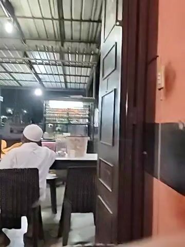 The video shows the interior of a modest outdoor restaurant space. A man in a white shirt and head covering sits at a dark wicker table and chair, facing away from the camera. Another man in a green shirt stands behind a food stall preparing food. There are a few other tables and chairs in the vicinity, and a display case stocked with drinks and snacks is also visible. The space has a corrugated metal roof with several light fixtures. The video appears to be filmed at night.