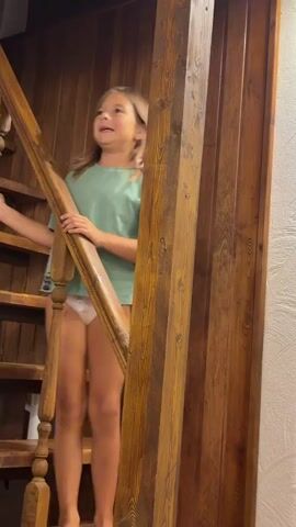A young girl stands on a wooden staircase, holding onto the banister. She is wearing a green t-shirt and white underwear. The staircase and surrounding walls are made of dark wood, suggesting an indoor setting, possibly a house or cabin. The lighting appears to be artificial, indicating it could be daytime or nighttime. The girl's expression is animated as she looks upwards, her mouth open as if speaking or singing. The scene is framed from a low angle, emphasizing the girl's presence on the stairs. There is no visible text in the media.