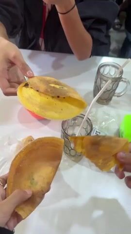 Multiple hands are visible around a white table. One hand on the left holds a golden-yellow folded pancake-like food item. Another hand in the center is tossing a similar golden-yellow food item, which is blurred due to motion. A third hand on the bottom right holds another folded golden-yellow food item. On the table, there are two clear patterned glasses each with a white straw, a small red lidded bowl containing a spoon, a clear plastic bag with more food, and a small green object. The upper torsos of two individuals in dark clothing are partially visible in the background, along with a dog's face on the far right.
