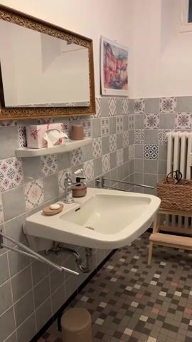 The video shows a small, tiled bathroom. On the left, there is a sink with a mirror above it. A tissue box and a small shelf with soap and a dispenser are on the shelf above the sink. To the right of the sink, there is a radiator with a wicker basket on top, and a small wooden step stool. Further to the right, a toilet is visible, and next to that is a blue bathtub with a shower curtain. The walls are covered in patterned tiles, and the floor is tiled in a mosaic pattern. The lighting is dim, and the camera moves from left to right, panning across the bathroom.