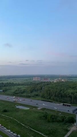 A high-angle, still shot shows a multi-lane highway cutting through a green, wooded area. Cars and trucks, including a large bus, move along the highway. In the distance are several large buildings under construction, with cranes visible. The sky is a gradient of light blue to pale pink at the horizon, suggesting dusk or dawn. To the sides of the highway, the land is covered in green grass and trees. Some parked cars are visible in the lower left corner of the shot.