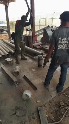 Two individuals are present in an outdoor, covered workshop area. The person on the left, wearing a dark outfit with orange accents and a cap, is using a tool to manipulate a cylindrical object on the ground. This object appears to be metal. The person on the right, wearing a dark t-shirt and jeans, stands observing, holding a tool in their right hand. The environment includes various metal pipes and structural elements, suggesting a fabrication or repair setting. The ground is concrete and appears somewhat weathered.