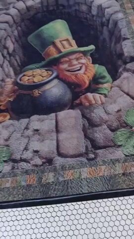 The video shows a close-up of a picture of a leprechaun smiling and clutching a pot of gold. The leprechaun is wearing a green hat and a green outfit. He has a red beard. The pot of gold is overflowing with gold coins. The picture is set against a stone background, and the bottom of the picture has a pattern of green and yellow rectangles. The video is shot from a slightly low angle. The floor beneath the picture is tiled with white hexagonal tiles.